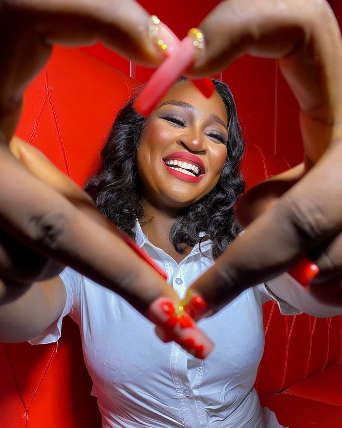 Smiling woman in white shirt making a heart shape with fingers against a red background.